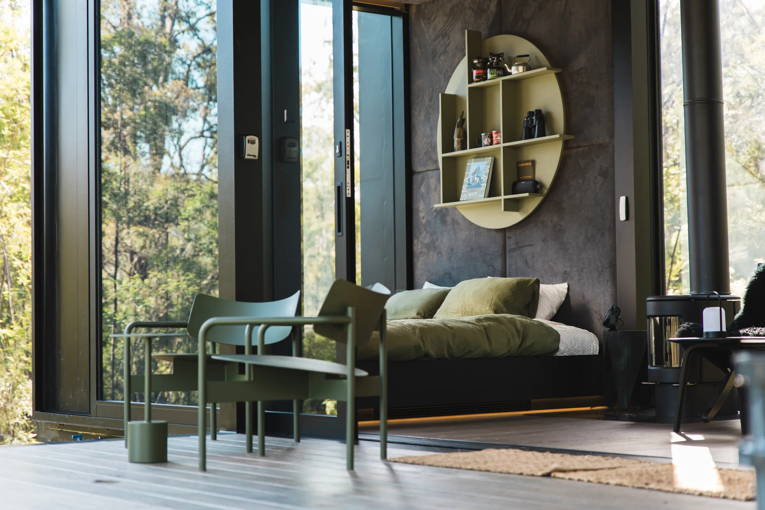 Modern bedroom with large glass doors, green armchairs, a bed with green bedding, and a circular wall shelf.