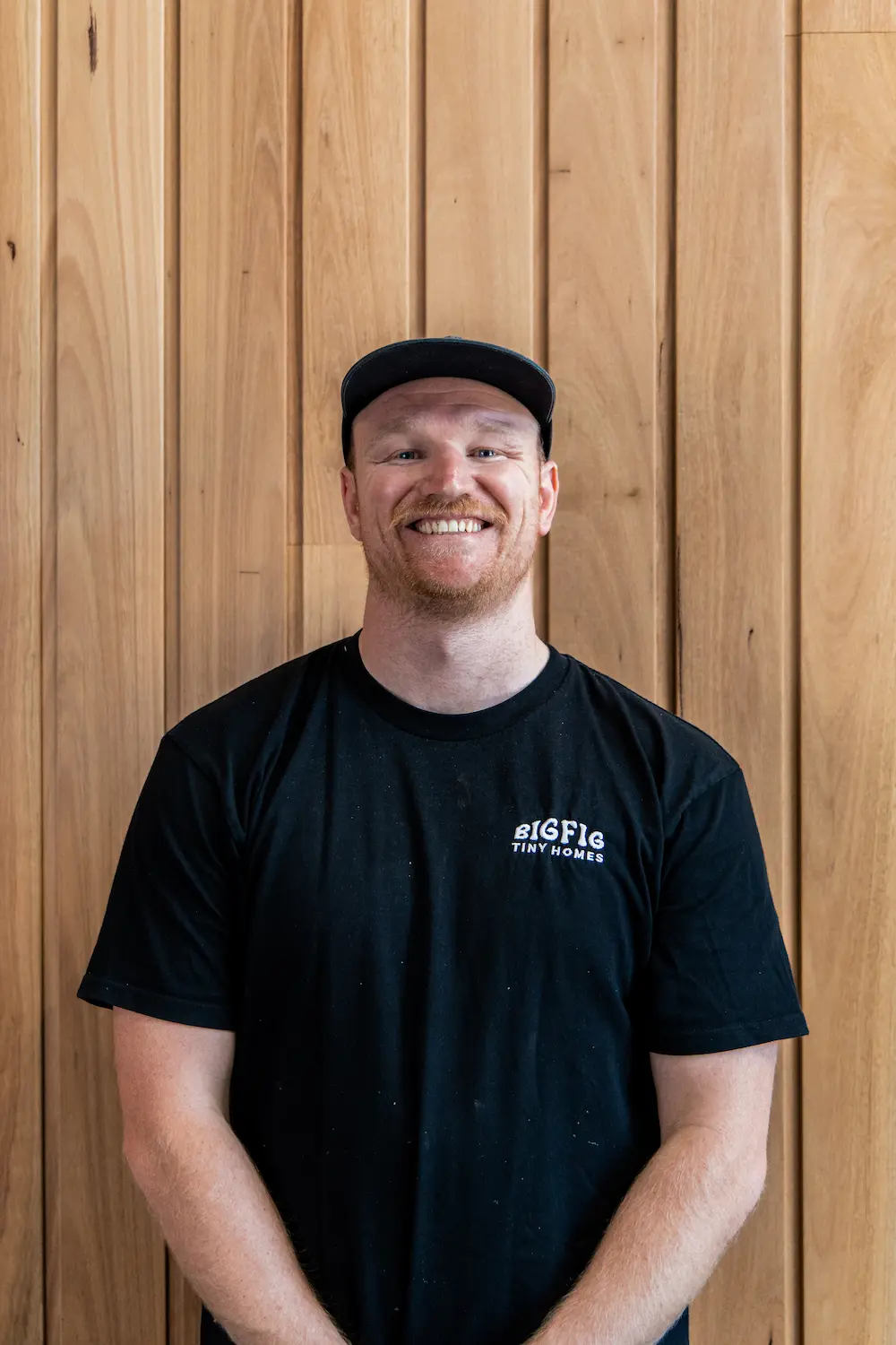 Smiling man with red beard wearing a black cap and black BigFig Tiny Homes t-shirt in front of wooden panel background.