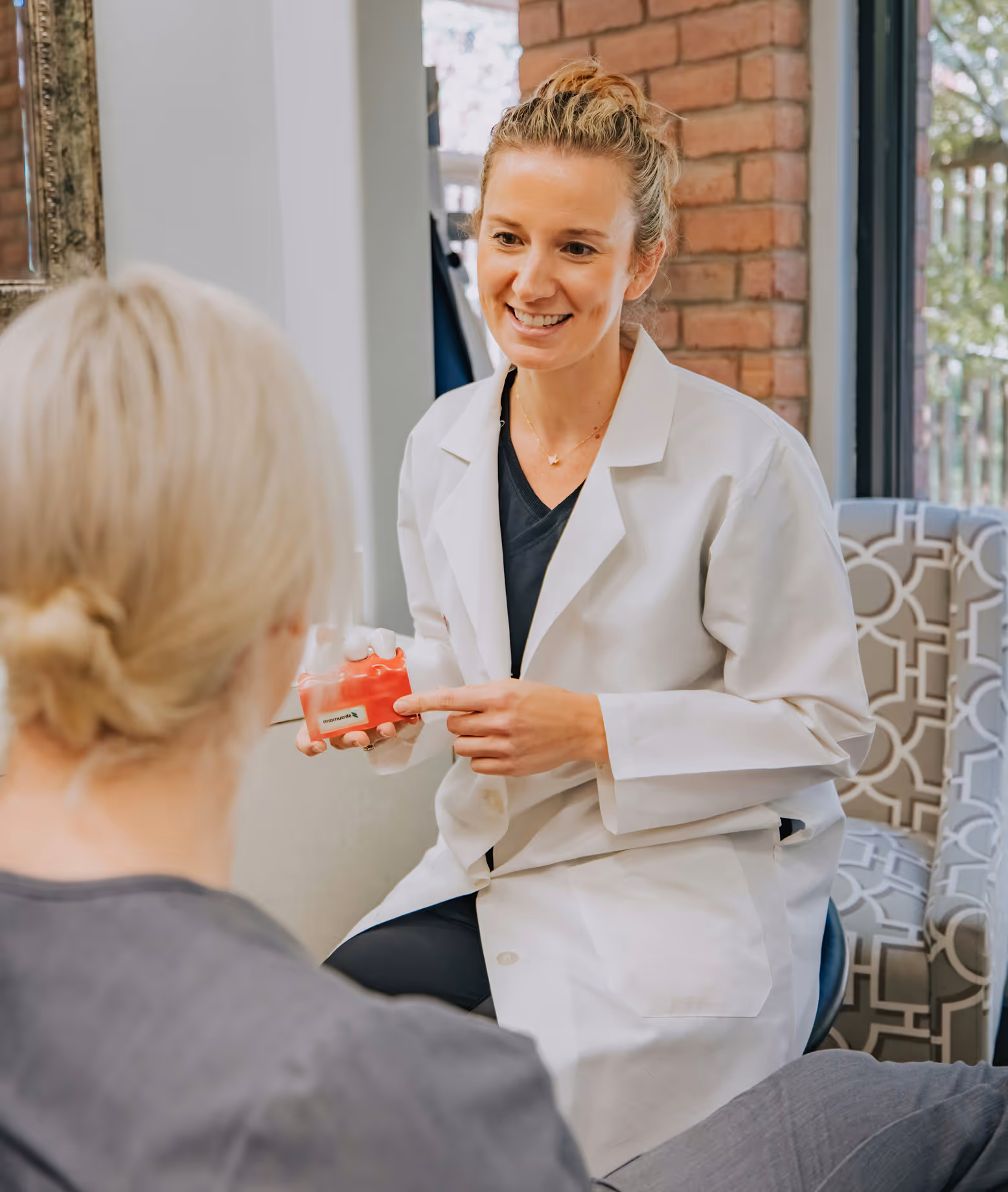 Smiling female dentist in a white coat showing a dental model to a seated patient.