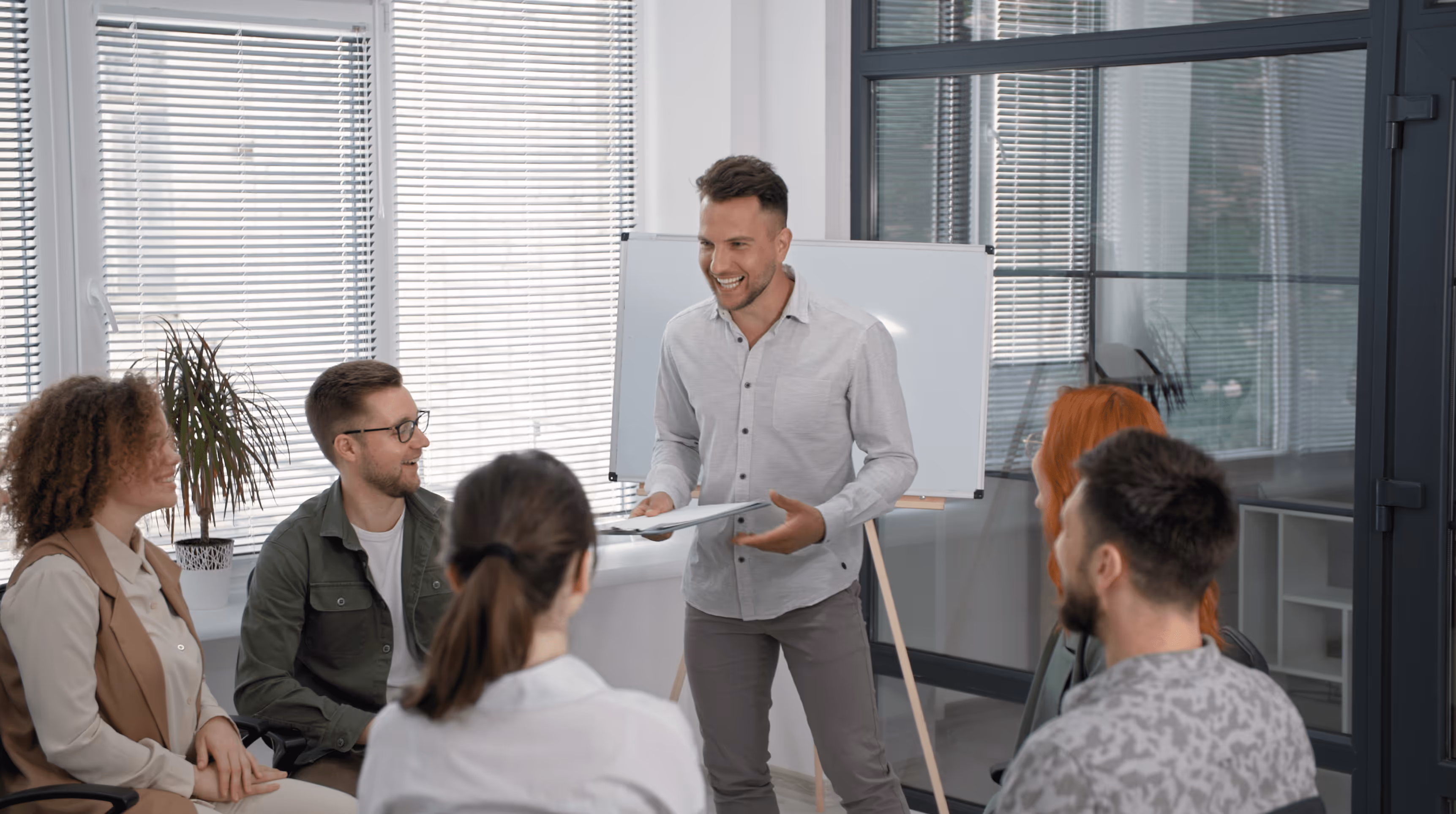 Man standing and presenting with a clipboard to four seated colleagues in a bright office with large windows.