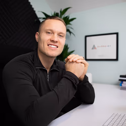 Smiling man with short blond hair wearing a black zip-up top, sitting at a desk with hands clasped, with a plant and framed picture in the background.