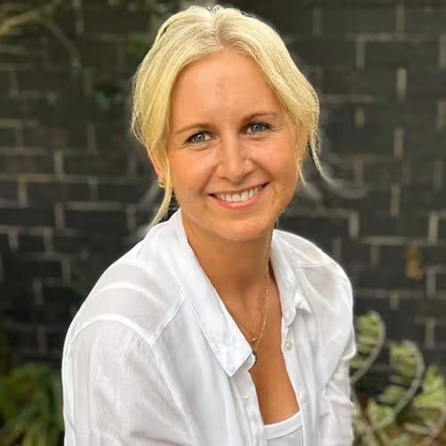 Smiling blonde woman wearing a white shirt posing outdoors with a dark brick wall background.