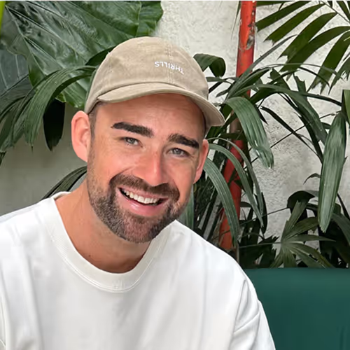 Smiling man with a beard wearing a beige cap and white sweatshirt sitting indoors near green plants.