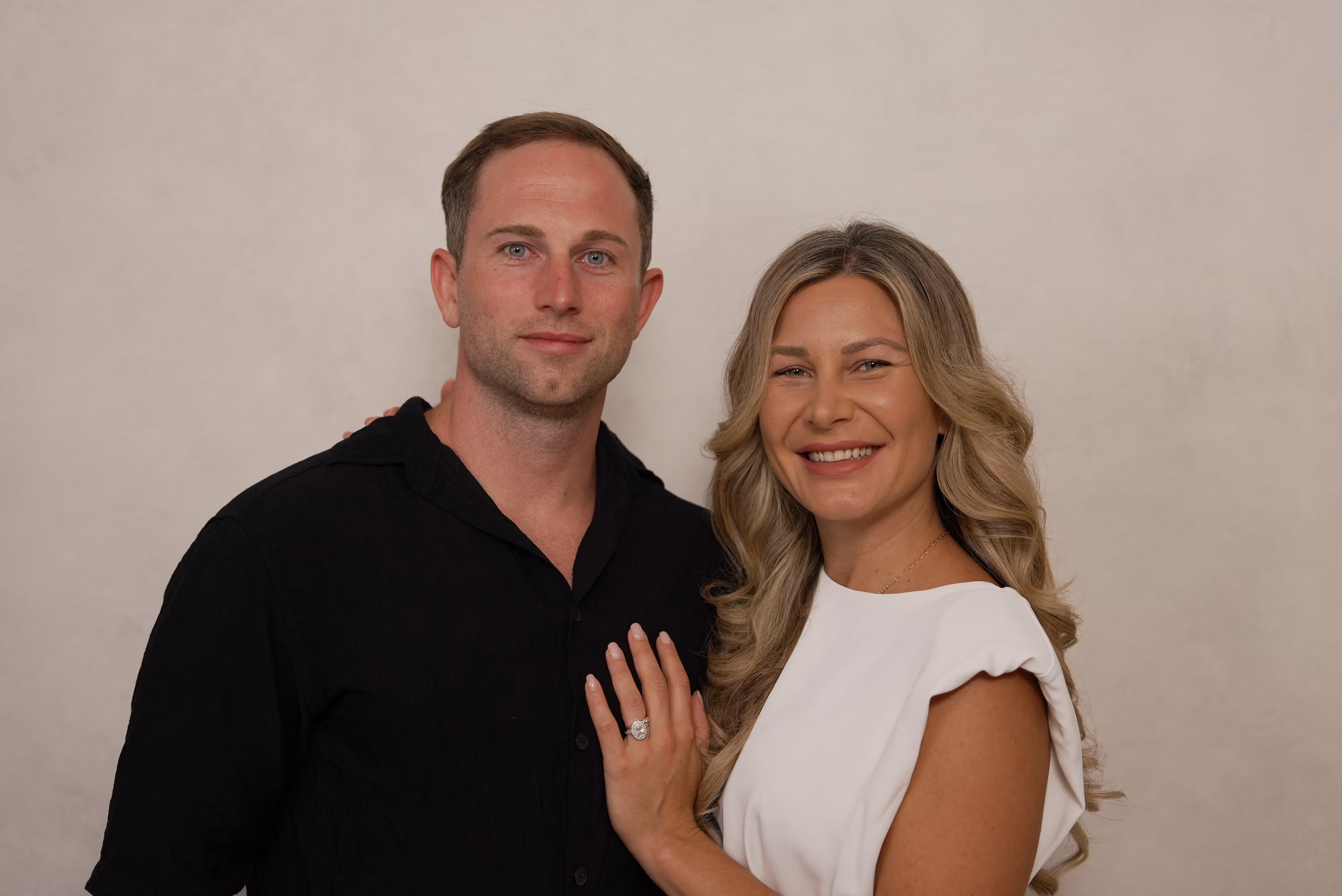 Smiling couple posing with the woman showing an engagement ring on her hand resting on the man's chest.