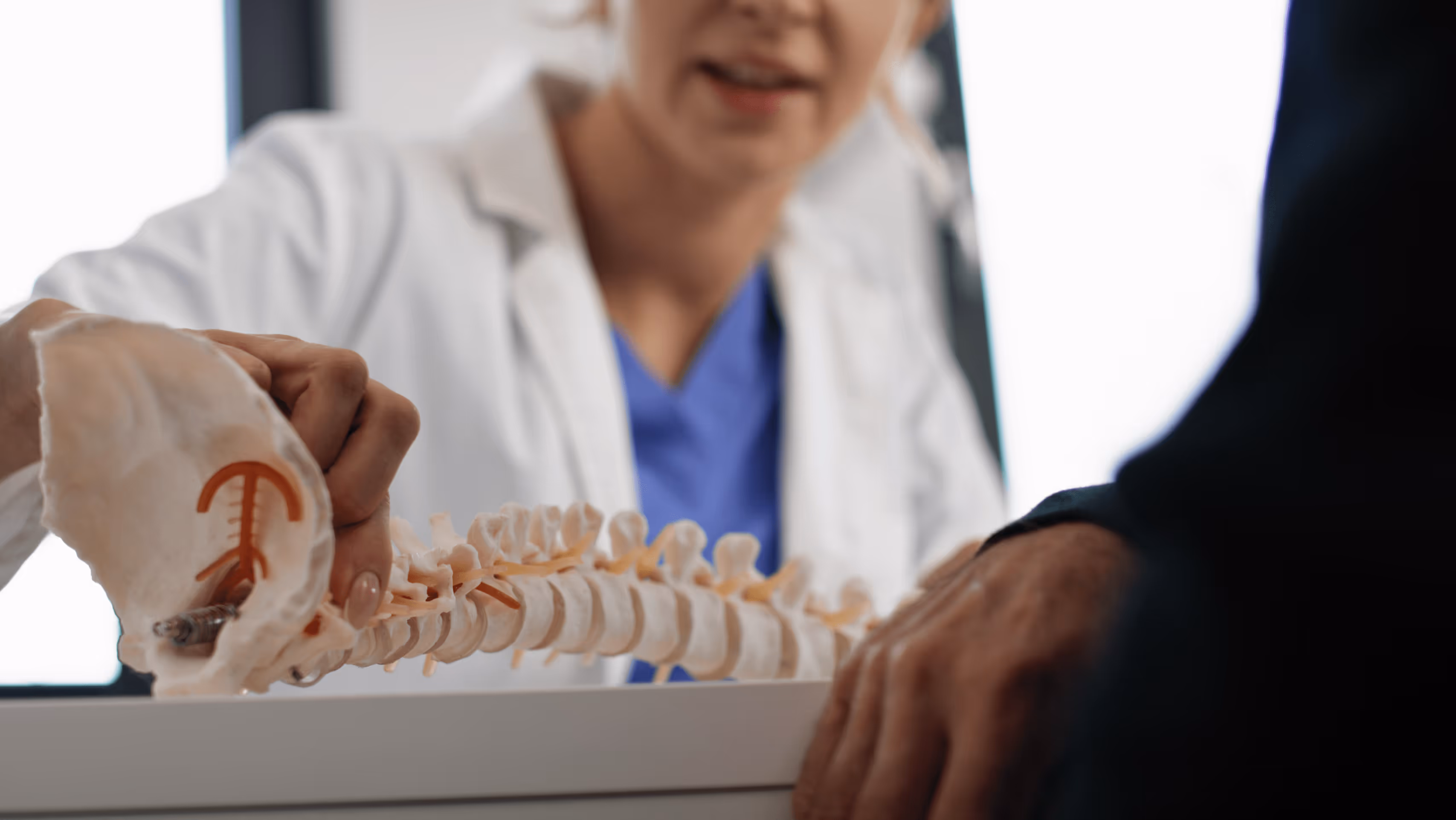 Medical professional in white coat holding a spinal model while explaining to a patient.