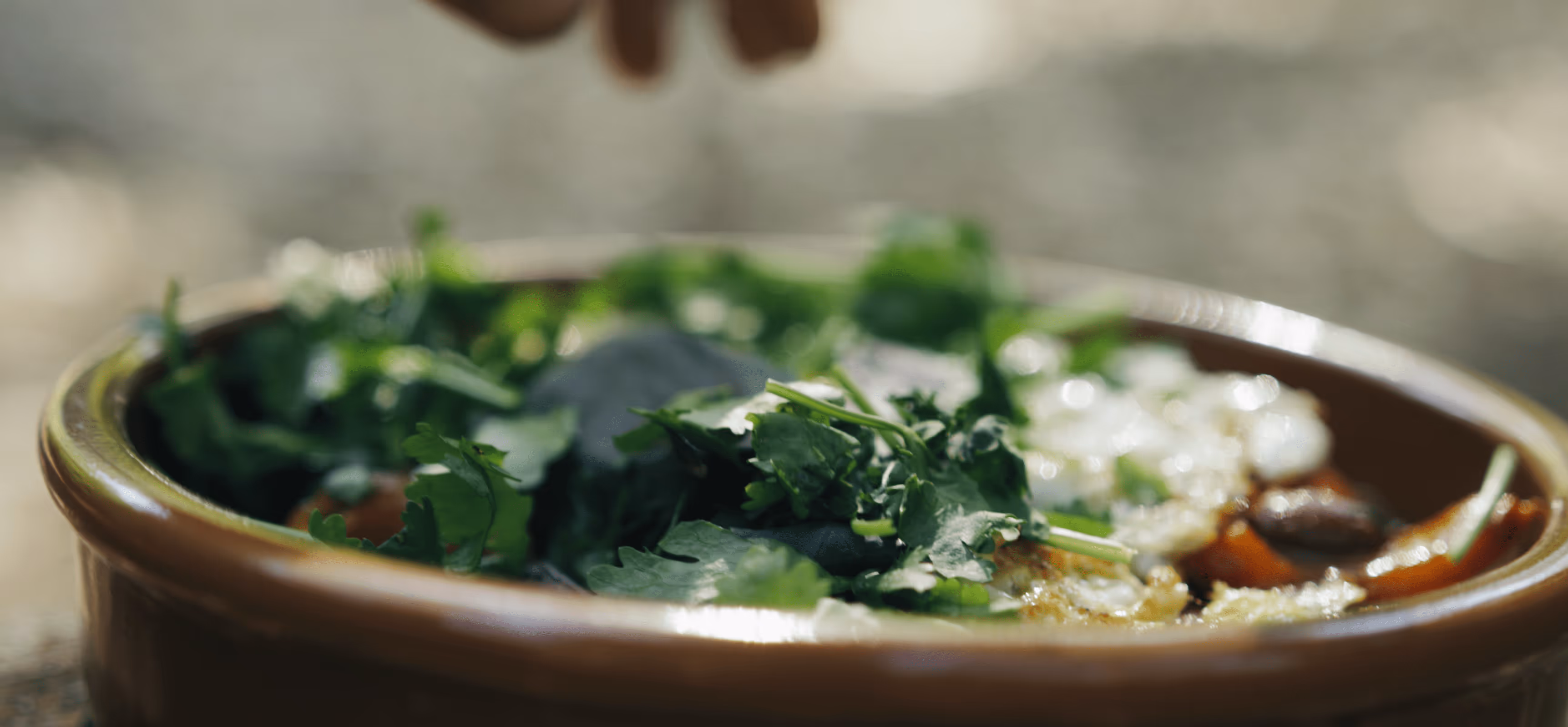 Close-up of fresh green herbs garnishing a dish in a brown bowl, with a hand poised above.