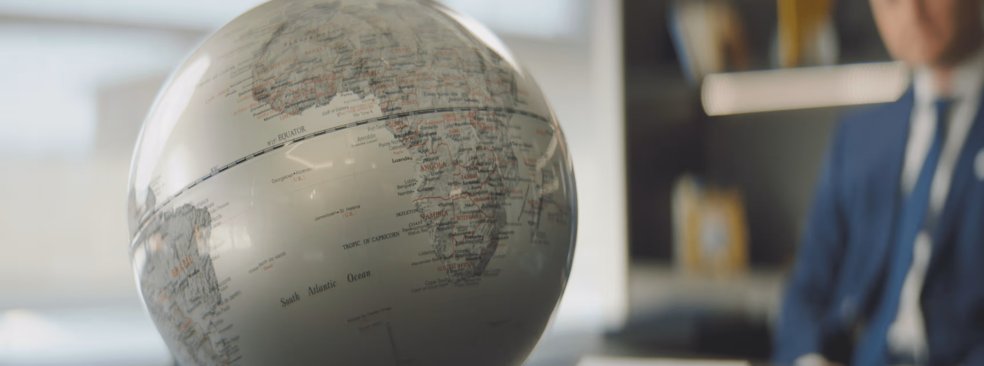 Close-up of a globe showing the South Atlantic Ocean, Africa, and Brazil with a blurred person in a suit in the background.