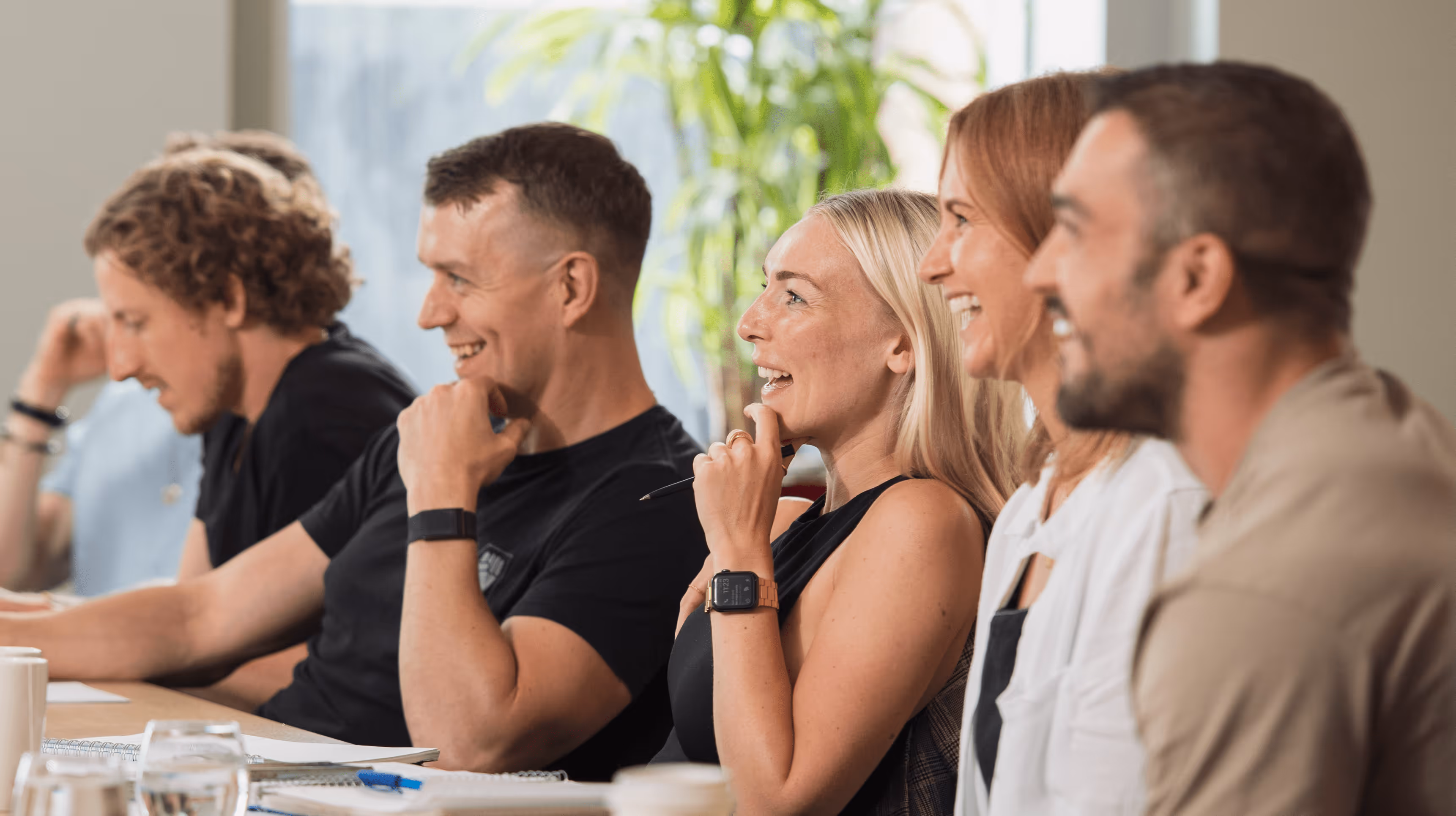 Group of five people sitting in a row at a table, engaged and smiling during a meeting or discussion.
