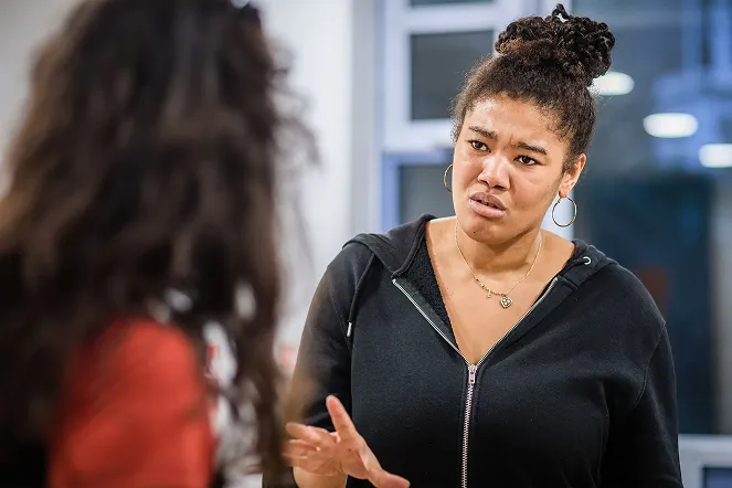 Woman with curly hair in a bun wearing a black hoodie gestures expressively while speaking to another person.
