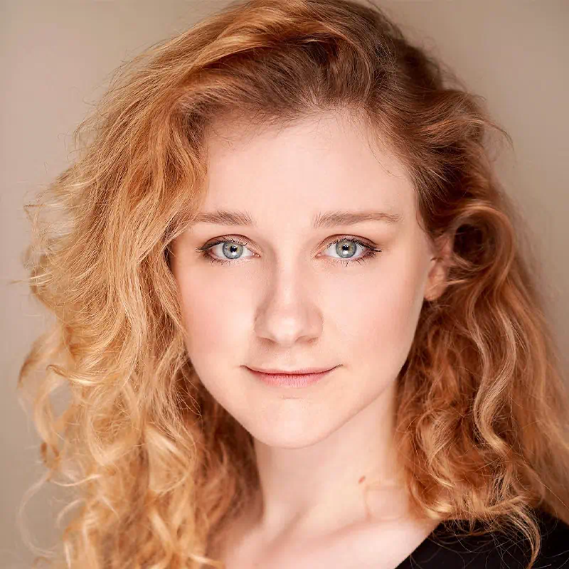 Close-up portrait of a young woman with curly red hair and blue eyes looking directly at the camera against a neutral background.