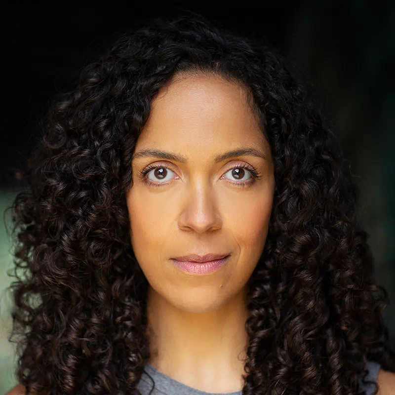 Woman with long curly dark hair and brown eyes looking directly at the camera against a dark background.