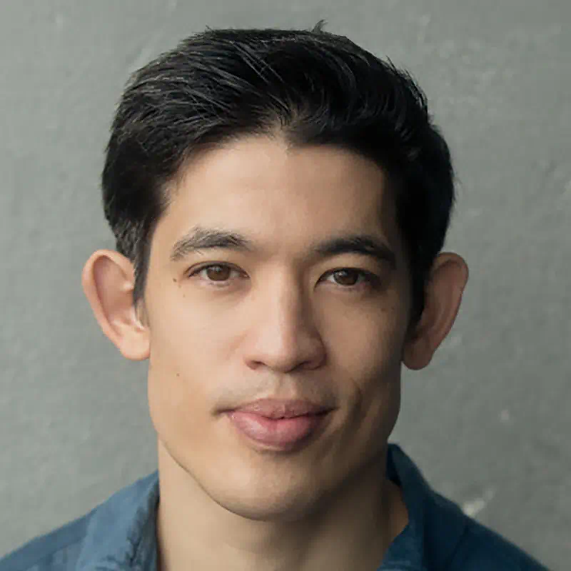 Headshot of a young Asian man with short black hair and a slight smile wearing a blue shirt, against a gray background.