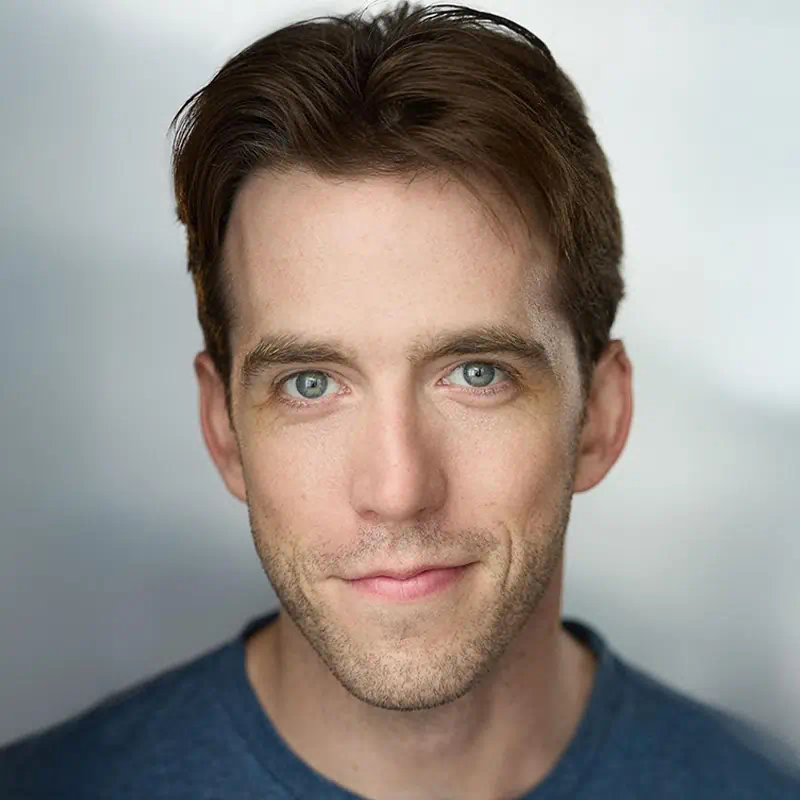 Close-up portrait of a young man with short brown hair, blue eyes, and light stubble, wearing a blue shirt and smiling softly.