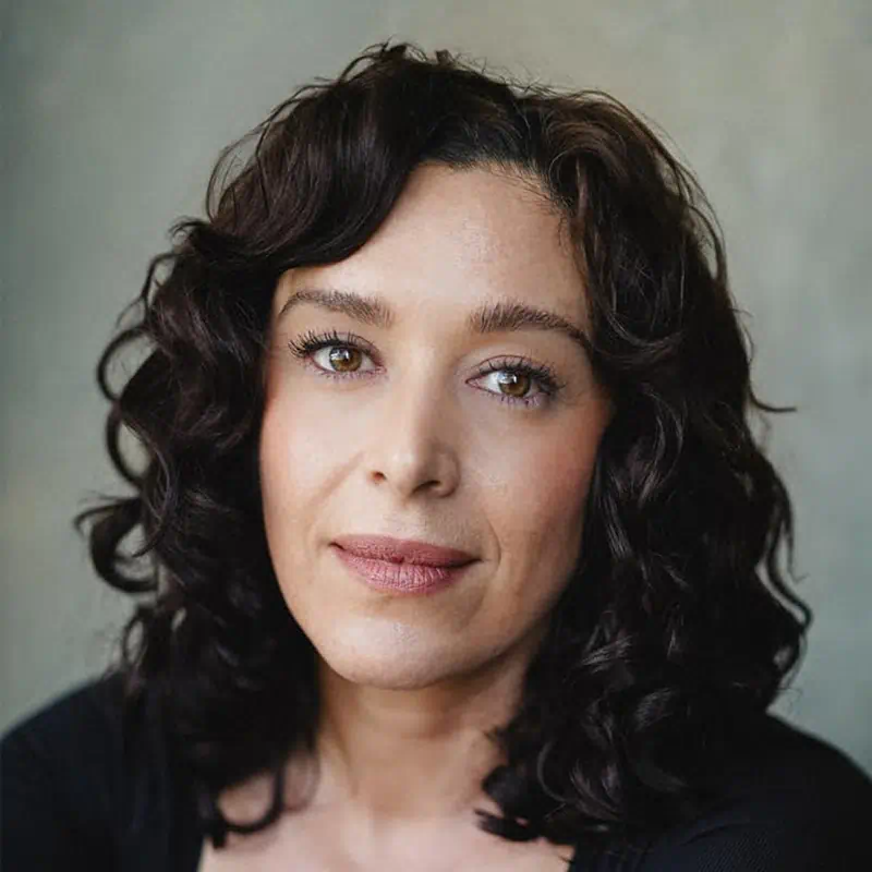 Portrait of a woman with shoulder-length curly dark hair and light makeup against a plain background.