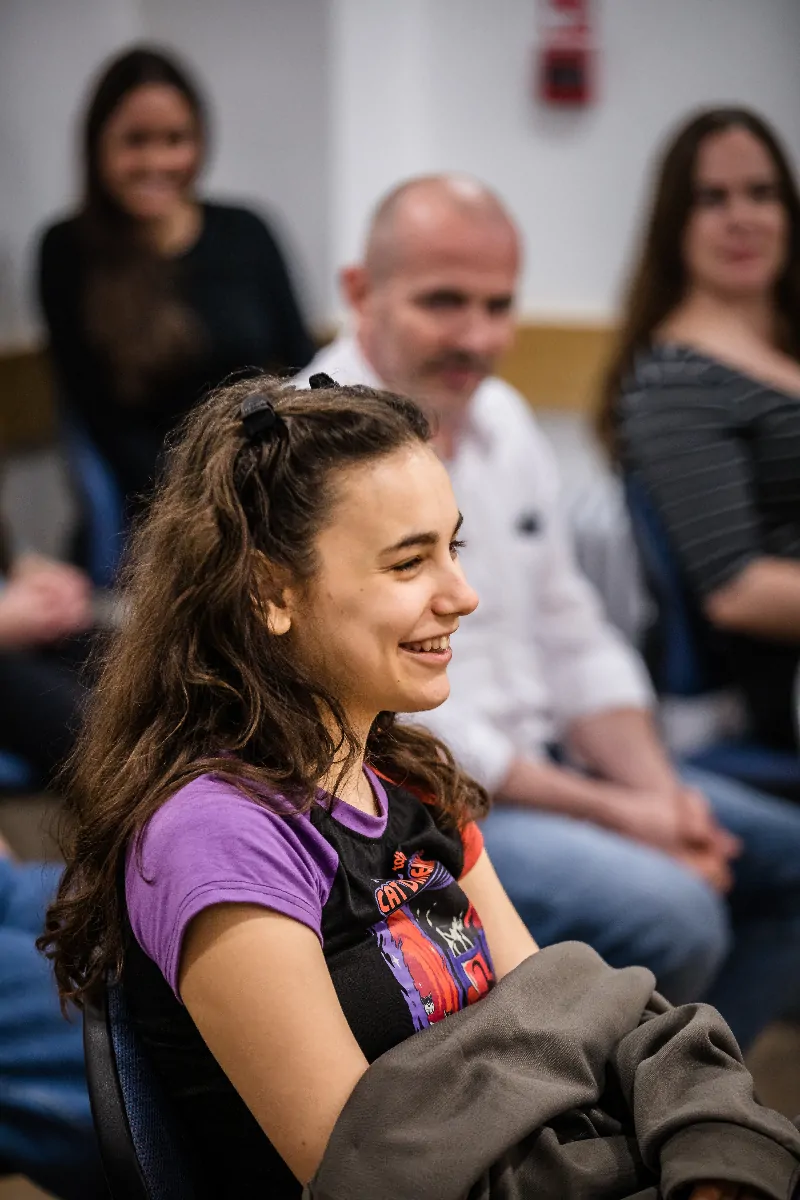 Young woman with long hair smiling and sitting in a group setting with three blurred people in the background.
