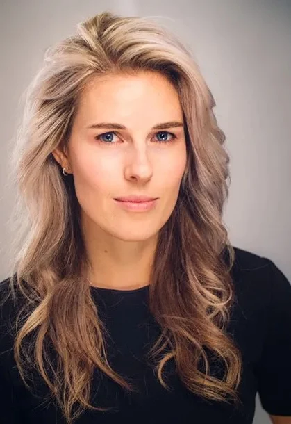 Portrait of a woman with long wavy blonde hair and blue eyes wearing a black top against a neutral background.