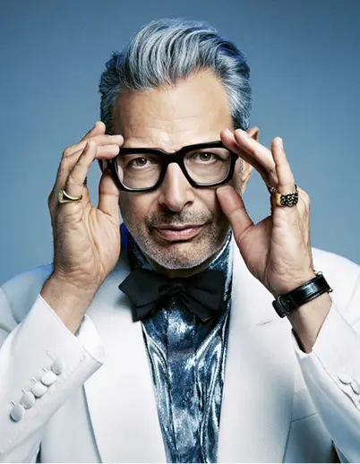 Man with gray hair wearing black glasses, a white suit jacket, a metallic shirt, and a black bow tie, adjusting his glasses with both hands.