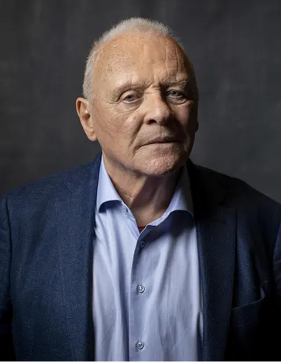 Portrait of an elderly man with short white hair wearing a blue suit jacket and light blue shirt against a dark background.