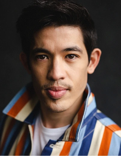 Close-up portrait of a young man with short dark hair, wearing a striped jacket with blue, orange, and white colors over a white shirt, looking directly at the camera against a dark background.