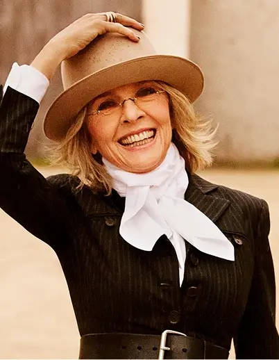Smiling woman wearing a tan hat, glasses, a black pinstripe jacket, a white scarf tied around her neck, and a wide black belt.