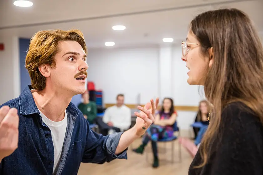 Young man with mustache and rolled-up sleeves gestures intensely while talking to a woman with glasses in a classroom with seated students in the background.