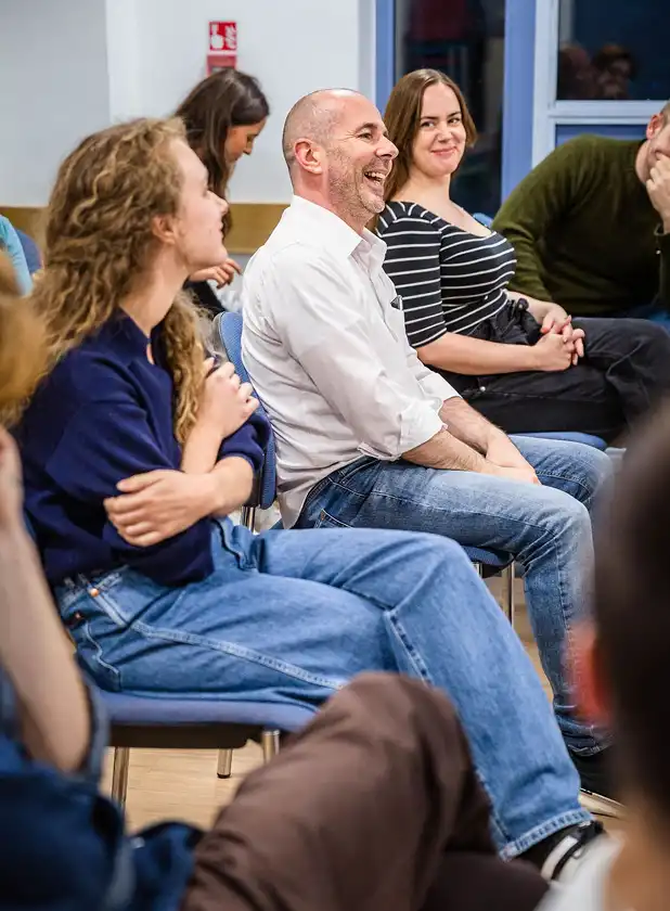 Group of adults sitting in chairs in a casual classroom setting, engaged and smiling during a discussion.