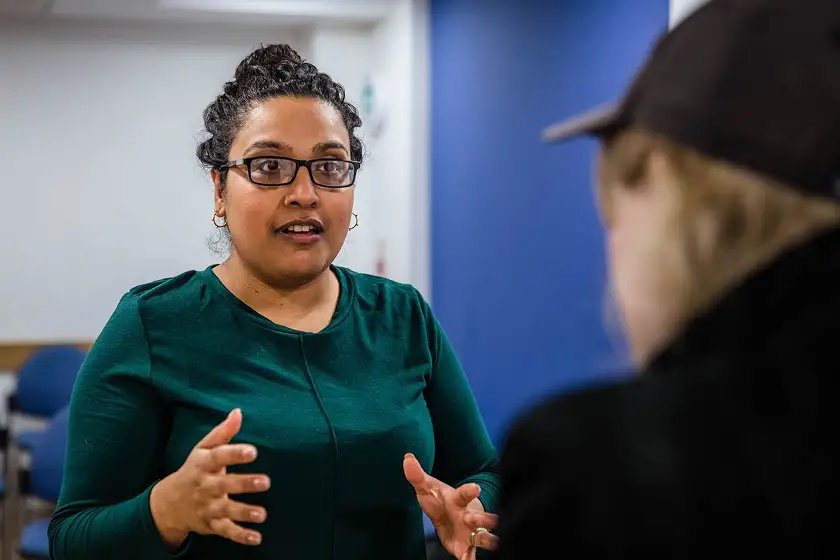 Woman with glasses and green shirt speaking and gesturing with hands to another person wearing a dark cap.