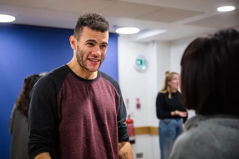 Man with short curly hair and a beard wearing a maroon and black shirt, talking and smiling indoors with blurred people in the background.