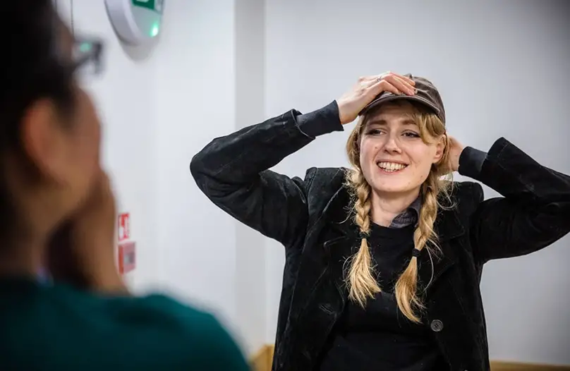 Smiling woman with braided blonde hair adjusting her dark cap during a conversation with another person.