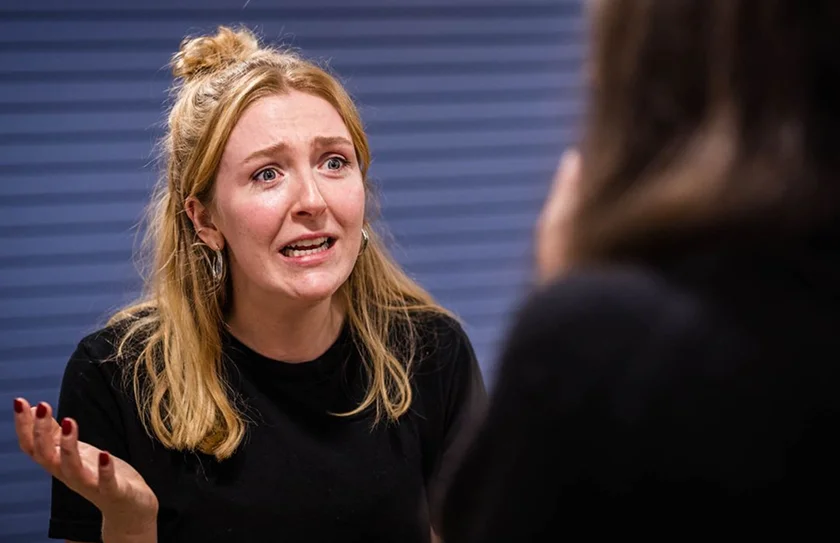 A woman with blonde hair and a black shirt gesturing with her hand while talking emotionally to another person.