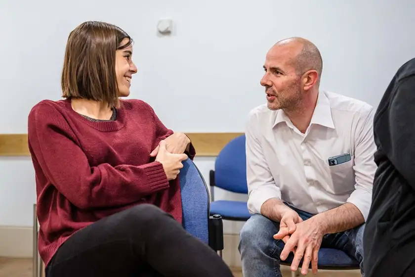 A man and a woman sitting on chairs engaged in a friendly conversation in a casual indoor setting.