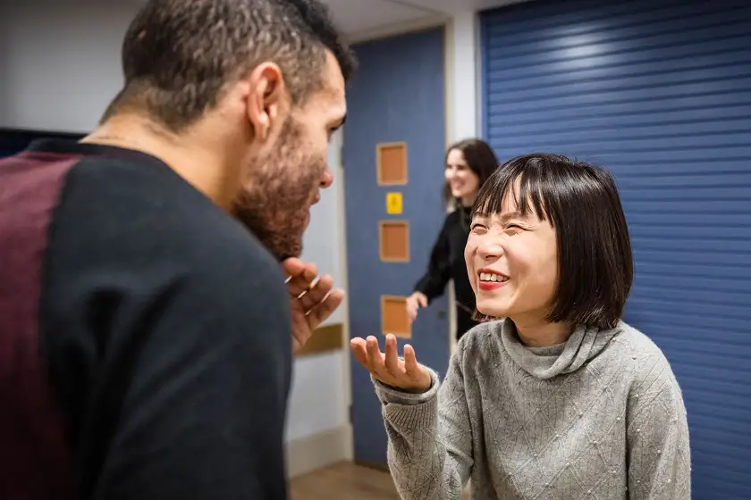 A woman in a gray sweater laughing and gesturing with her hand while talking to a man with a beard in an indoor setting, with another woman smiling in the background.