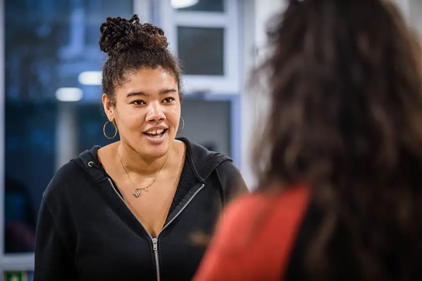 Young woman with curly hair tied up, wearing hoop earrings and a necklace, speaking to another person in an indoor setting.