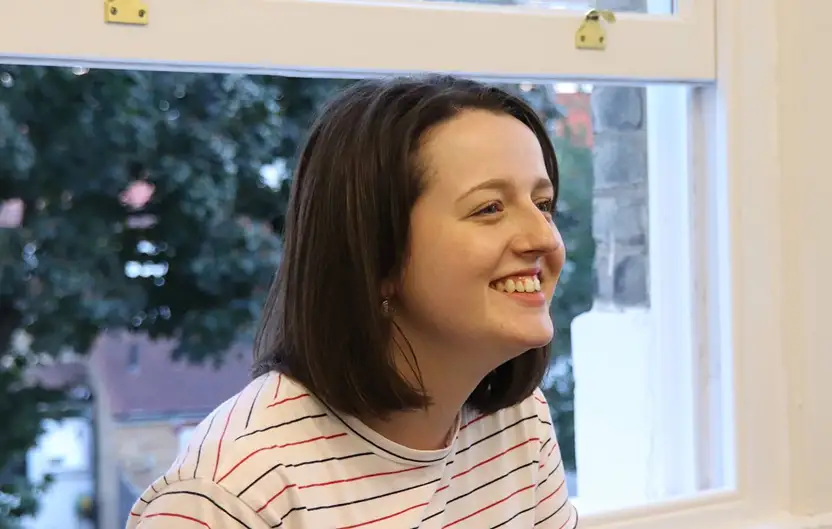 Smiling woman with shoulder-length brown hair sitting near a window with a blurred outdoor background.