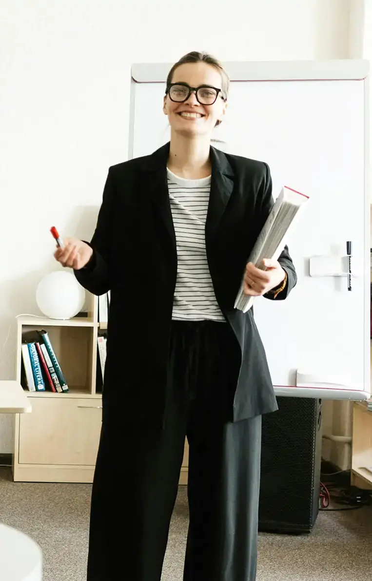 Smiling woman wearing glasses and a black blazer holding a marker and a rolled-up newspaper standing in front of a whiteboard.