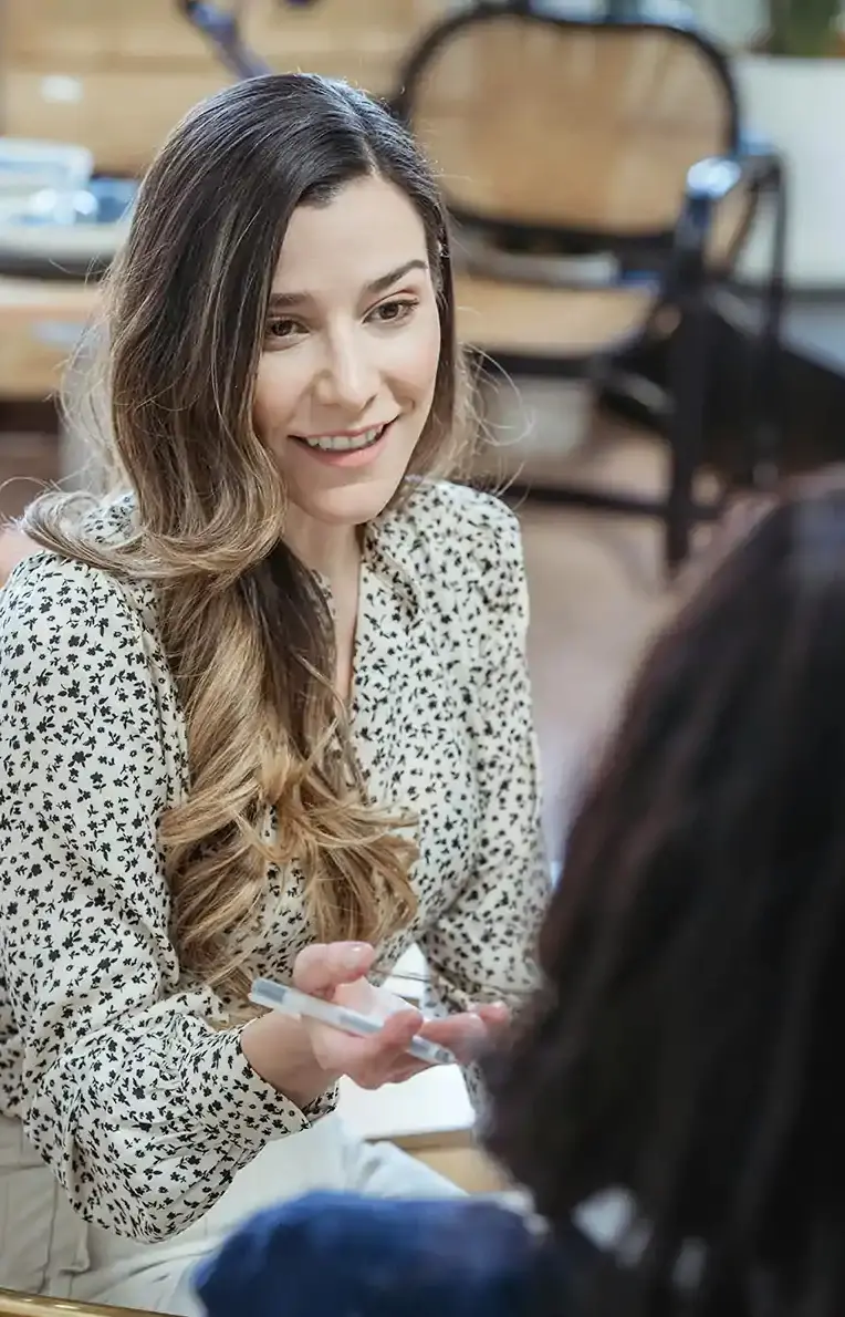 Smiling woman with long wavy hair and a floral blouse holding a pen, engaged in conversation with another person.