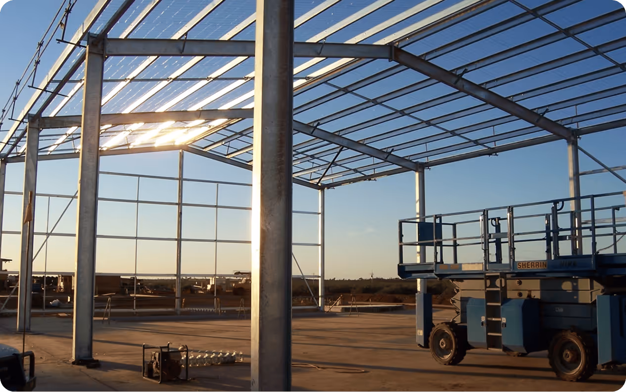 Steel frame structure under construction with a scissor lift on a concrete floor under a clear sky.