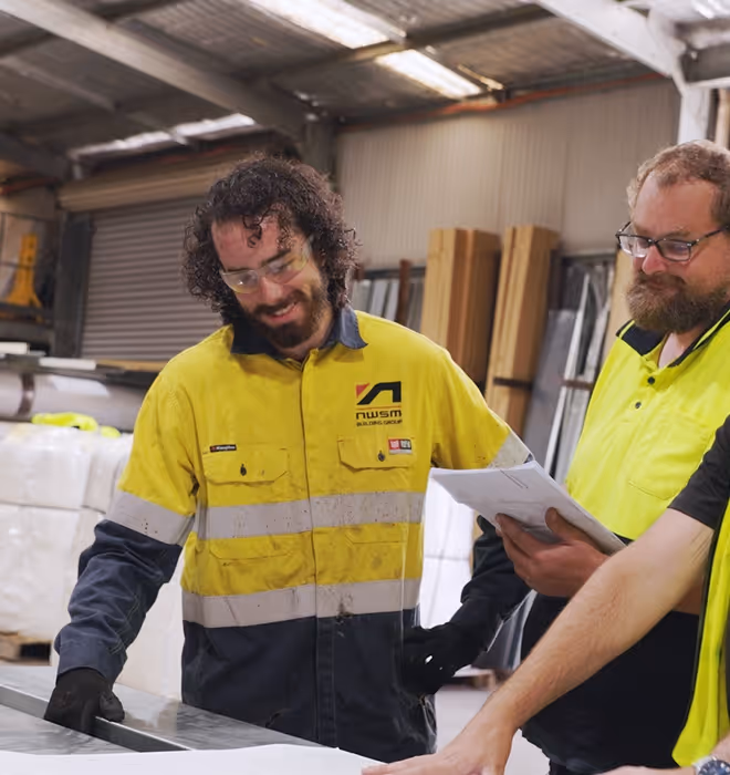 Two construction workers in high-visibility shirts reviewing plans inside a workshop.