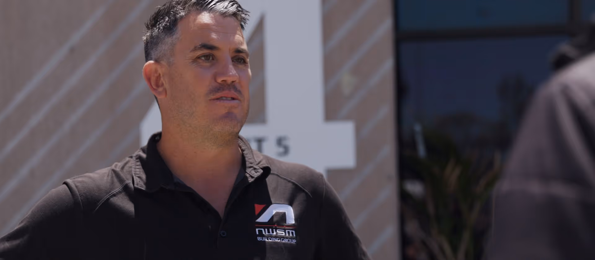Man wearing black NWSM Building Group polo shirt standing outdoors near a building.