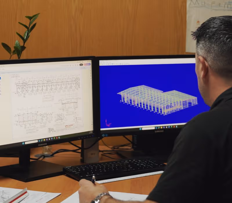 Man reviewing architectural blueprints and 3D building model on dual monitors at a desk.