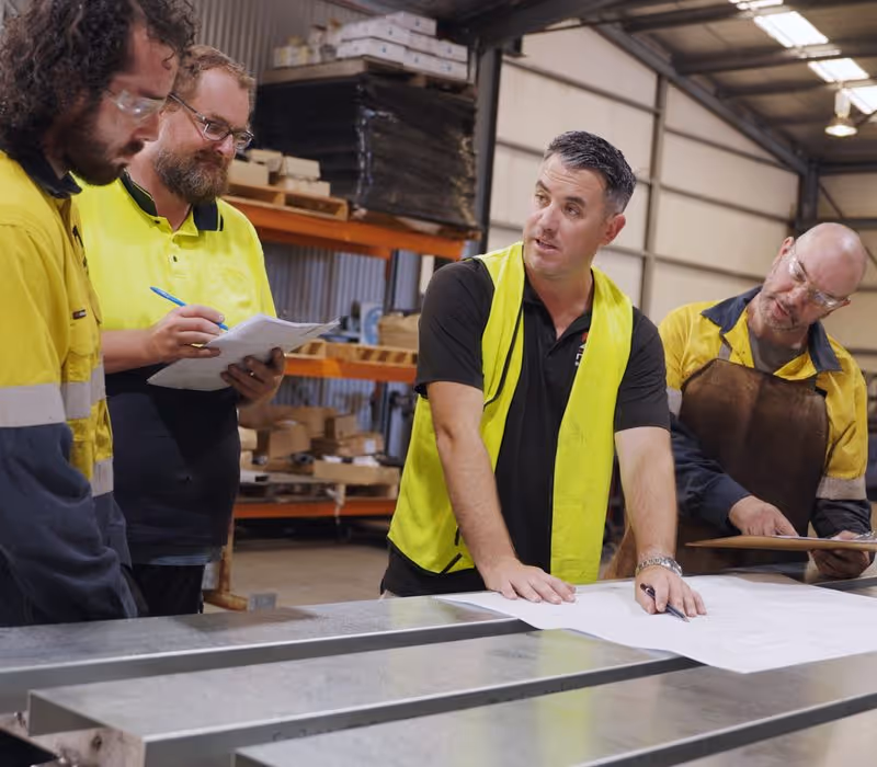Four workers in safety gear reviewing documents over a metal table in a warehouse.