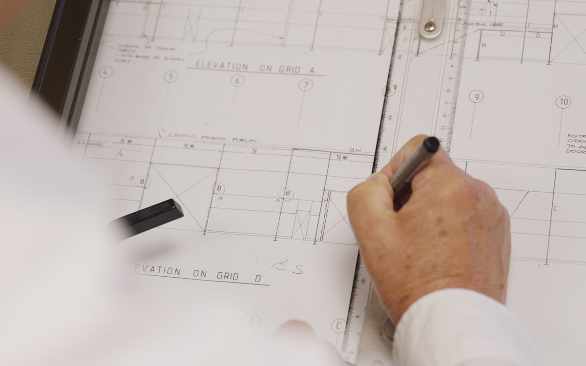 Close-up of a hand drawing architectural plans using a pen and ruler on drafted blueprints.