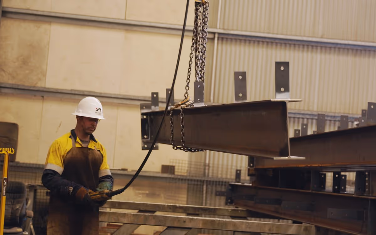 Worker in safety helmet and gloves operating crane to lift a large steel beam in a warehouse.