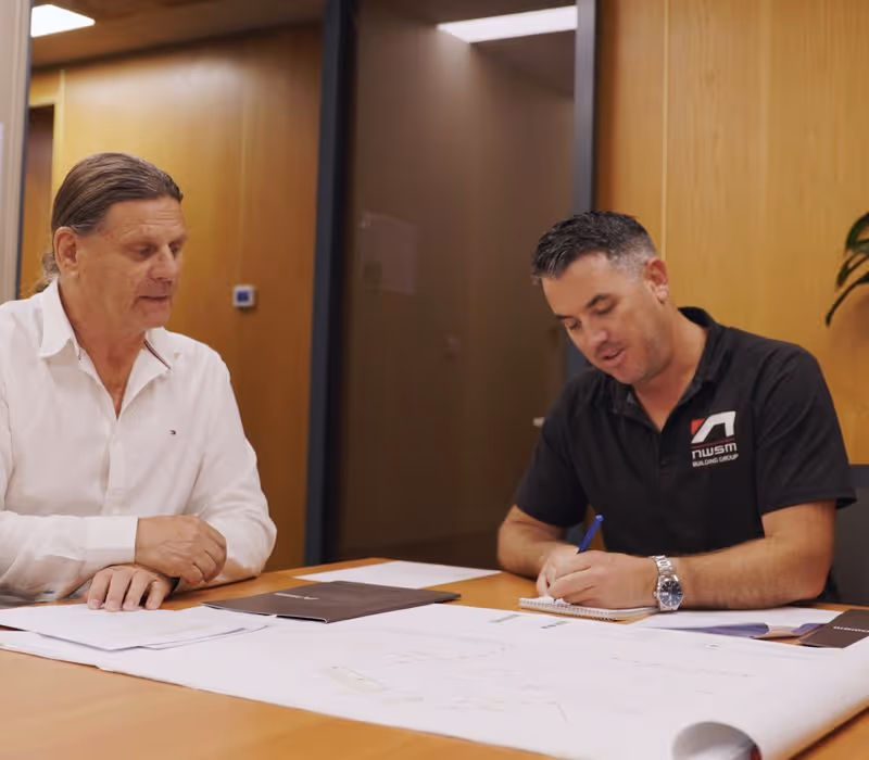 Two men at a table reviewing blueprints and documents during a meeting in an office.