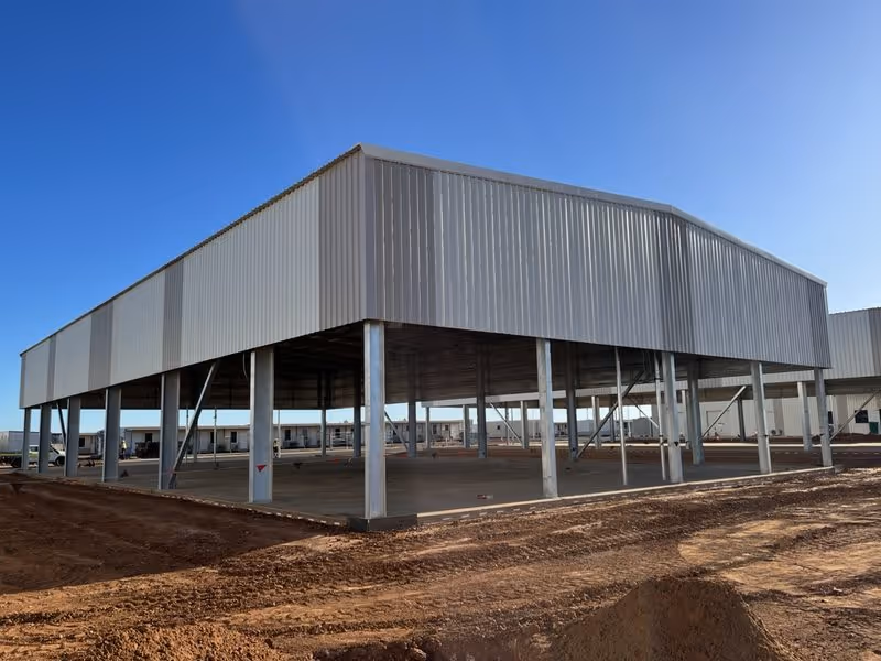 Steel frame structure under construction on a cleared dirt lot under a clear blue sky.