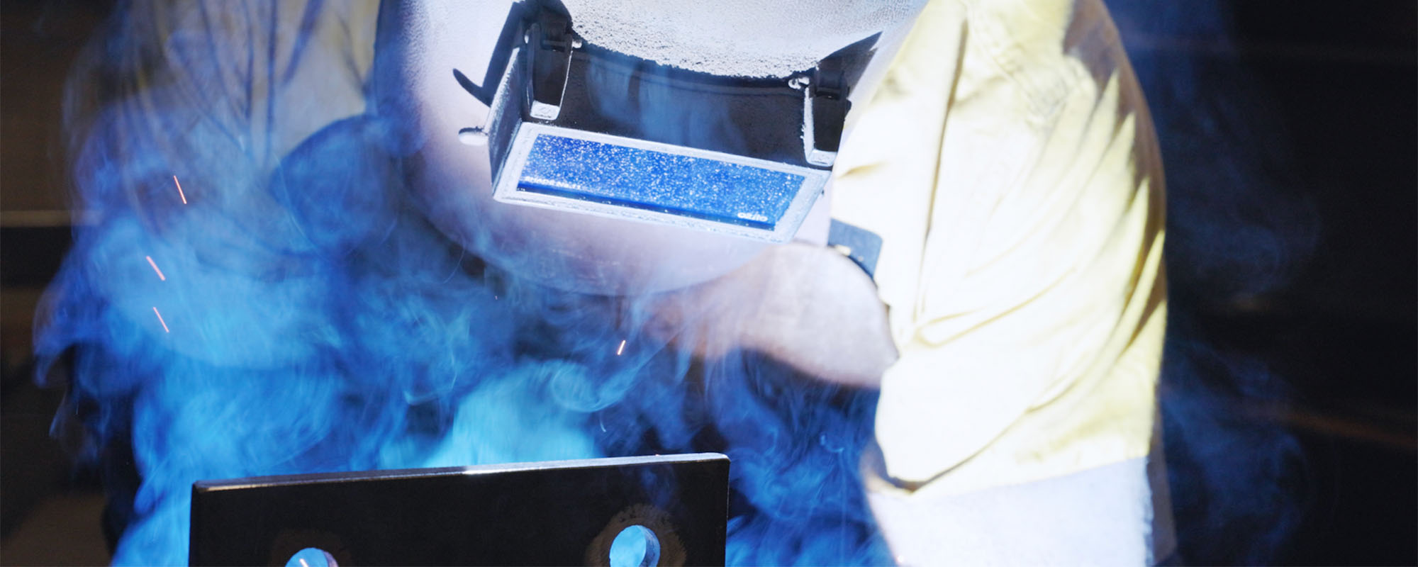 Welder wearing protective helmet and gloves working with bright blue sparks and smoke.