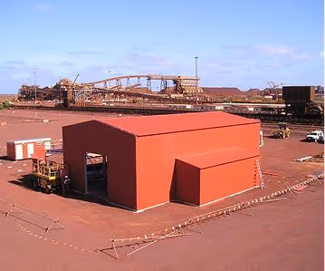 Red industrial building on reddish ground with large conveyor system and machinery in the background.