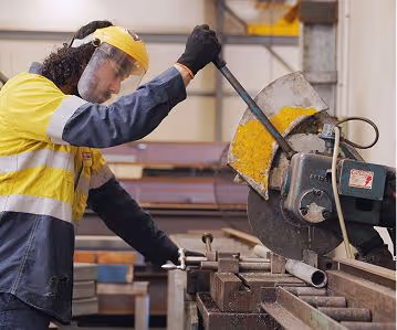 Worker wearing safety gear operating an industrial metal cutting saw on a metal pipe.