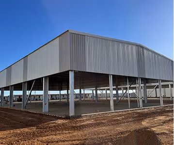 Metal industrial building under construction with steel frame and partial paneling on dirt ground.
