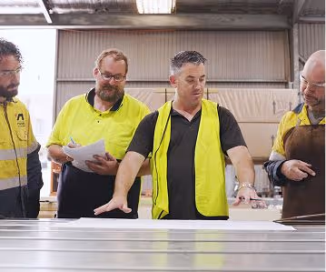 Four men in high-visibility workwear discuss and inspect metal sheets in a workshop.